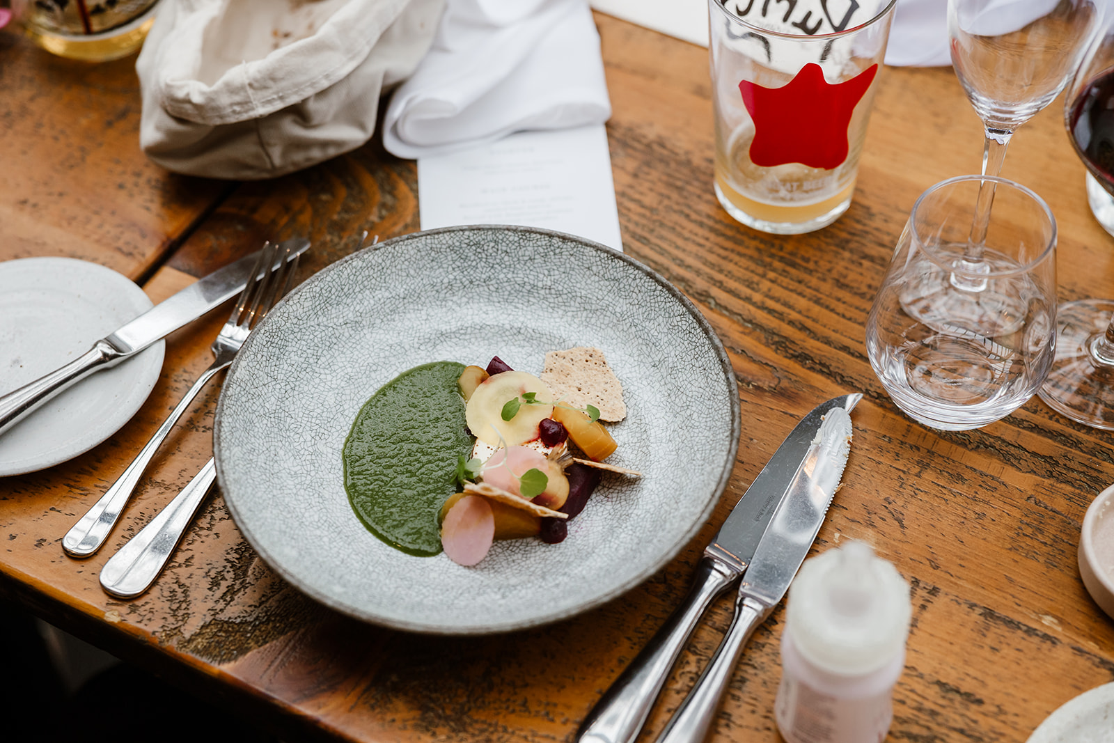 A plated dish with green purée, sliced vegetables, and a crisp garnish sits on a wooden table set with cutlery, glasses, a serviette, and a partially filled glass.