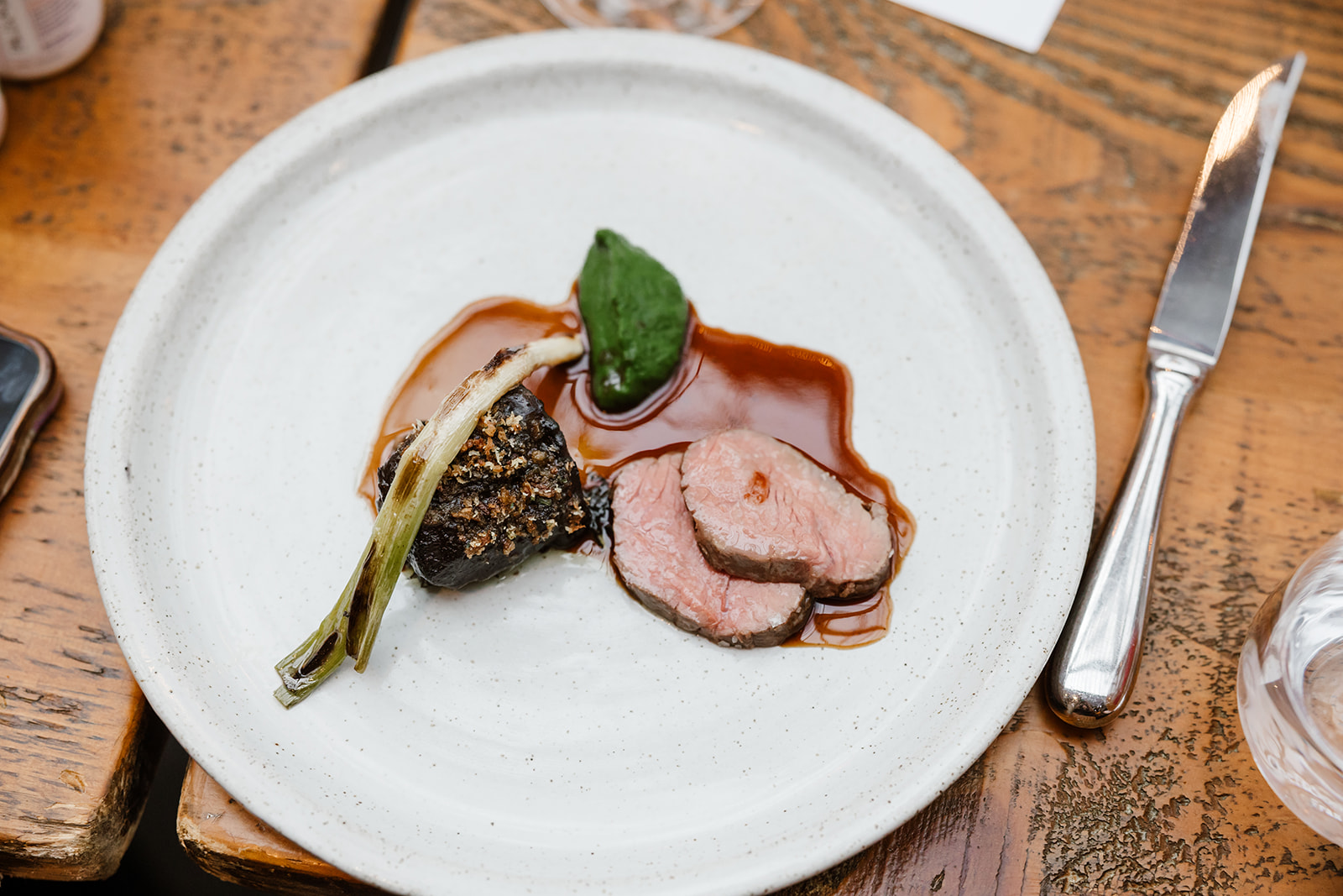 A white plate with two slices of cooked meat, a grilled spring onion, a dark vegetable topped with seeds, green leaf, and brown sauce, set on a wooden table with cutlery.