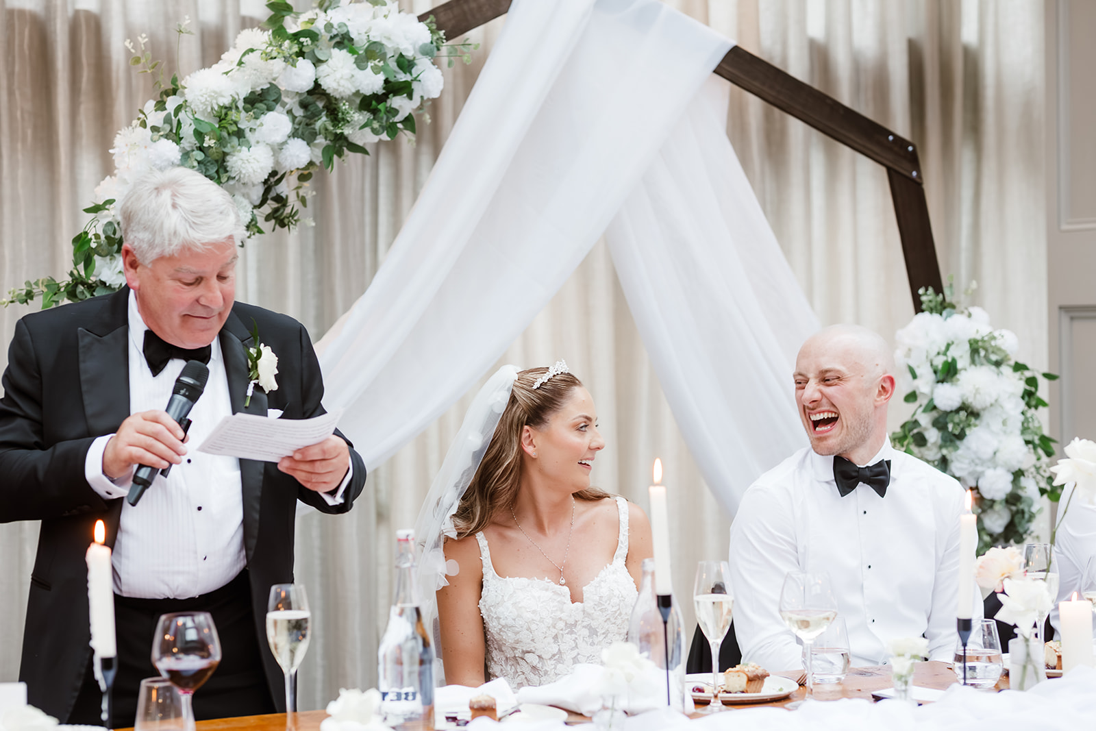 A man in a dinner jacket, the father of the bride, reads from a piece of paper at a wedding table as the smiling bride and groom sit beside him. The table is beautifully decorated with flowers and candles.