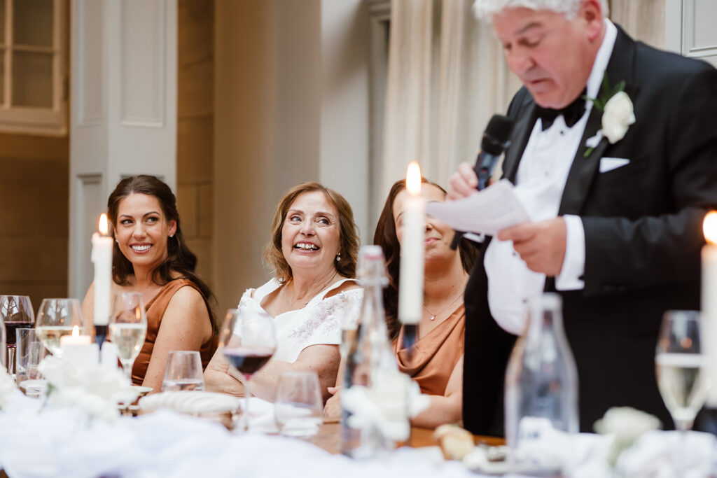 An older man in a dinner jacket, the father of the bride, gives the speech with a microphone at a table; women seated nearby listen and smile during an indoor event with candles and drinks.