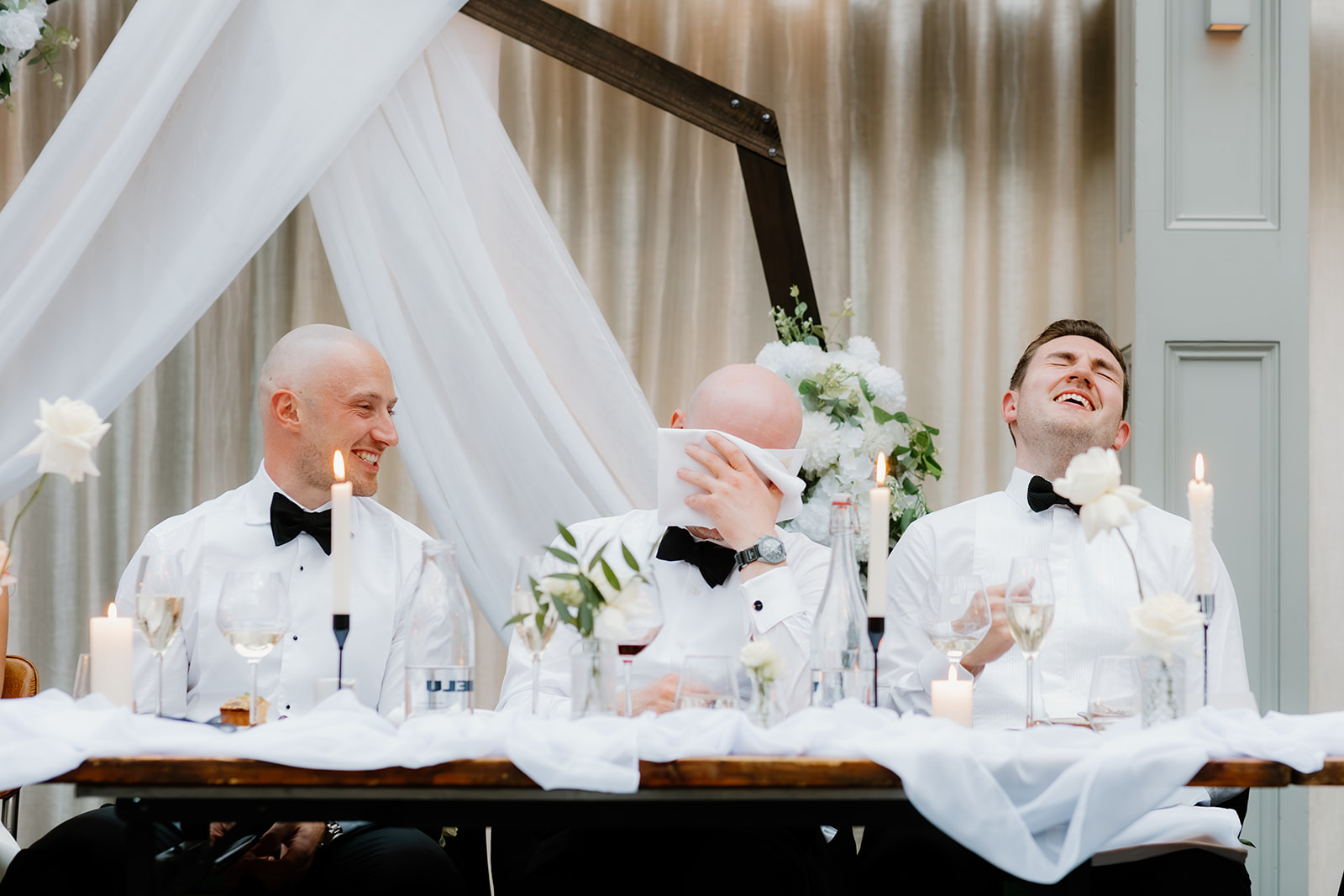 Three men in white shirts and black bow ties, including the usher, sit at a decorated table laughing; one wipes his face with a serviette. White drapery and candles set an elegant scene.