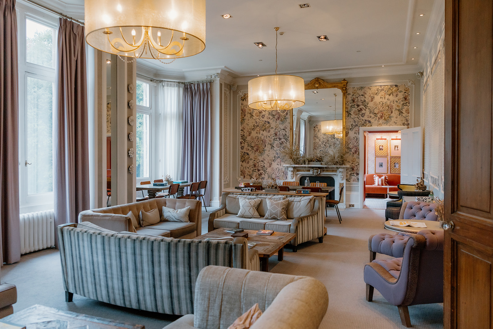 Elegant lounge with sofas, armchairs, coffee tables, large windows with curtains, chandeliers, a fireplace, and floral wallpaper. A large mirror hangs above the mantelpiece. Hampton Manor