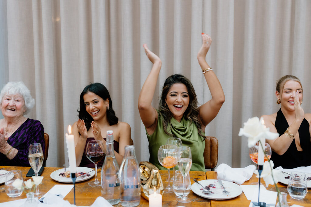 Four women seated at a dining table applaud and smile, with empty plates, glasses, and candles in front of them.