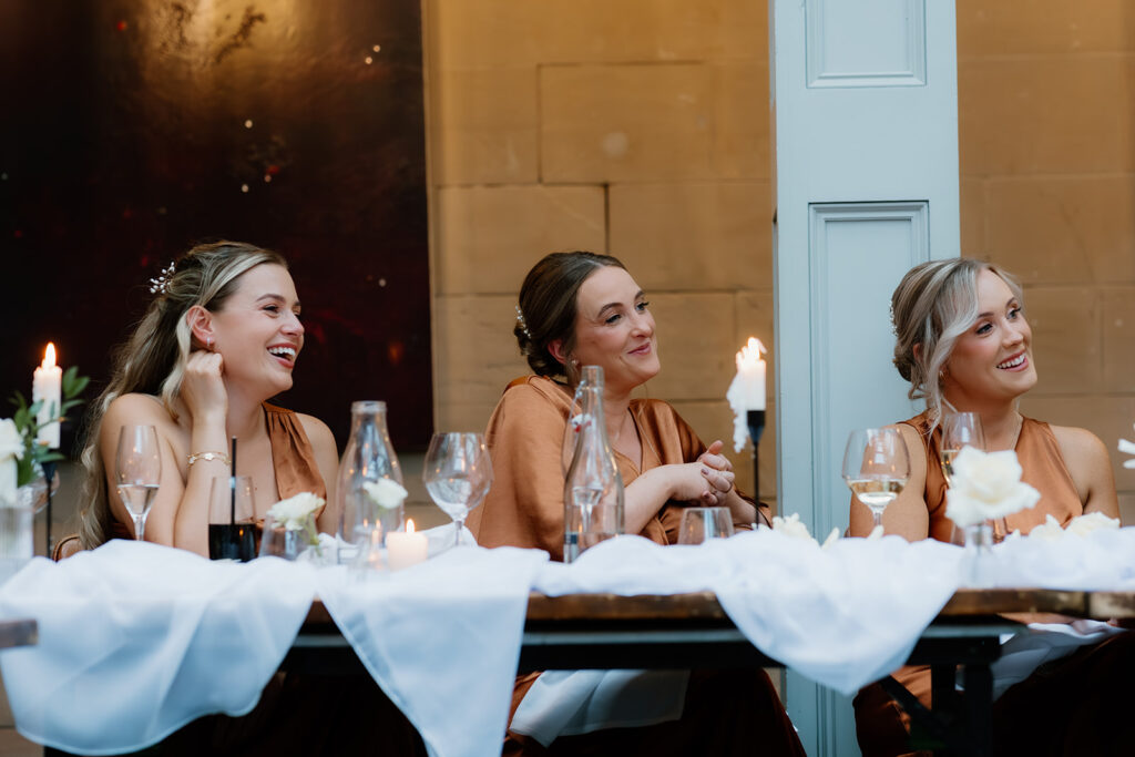 Three women in matching brown dresses sit at a decorated table with candles and drinks, smiling and listening attentively during an event.