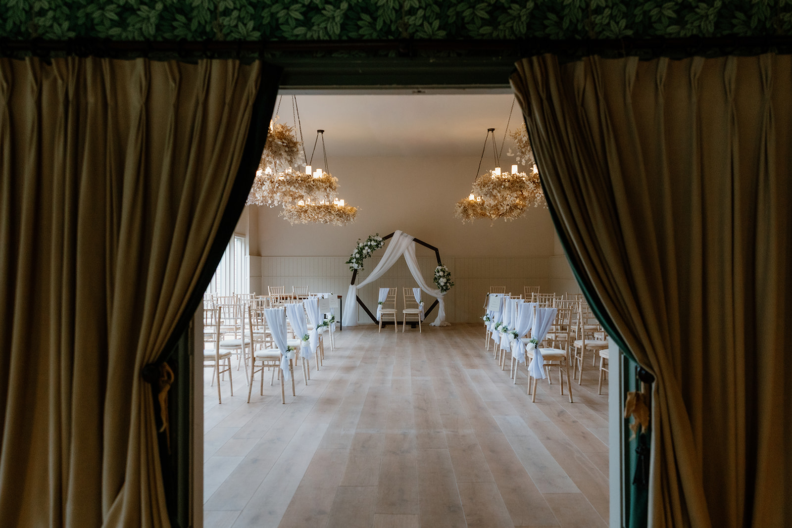A view through parted curtains reveals the Birches ceremony room, showcasing rows of chairs, elegant white decorations, and a beautiful floral arch at the front for an indoor wedding celebration.