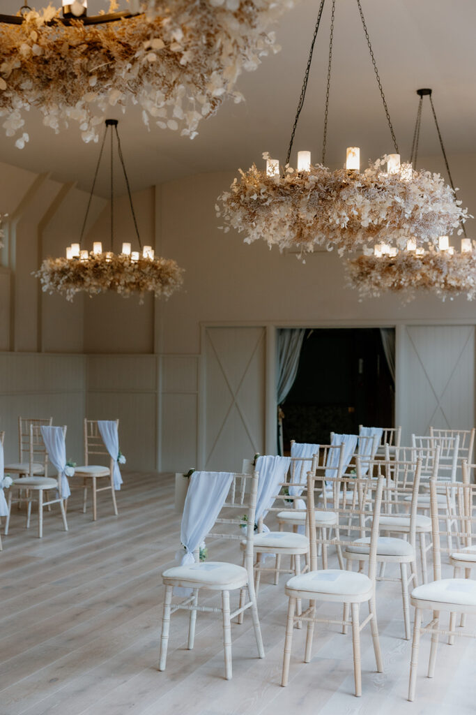 Rows of white chairs with draped fabric are arranged in the spacious, light-filled Hampton Manor ceremony room Birches, featuring wooden floors and large floral chandeliers overhead.