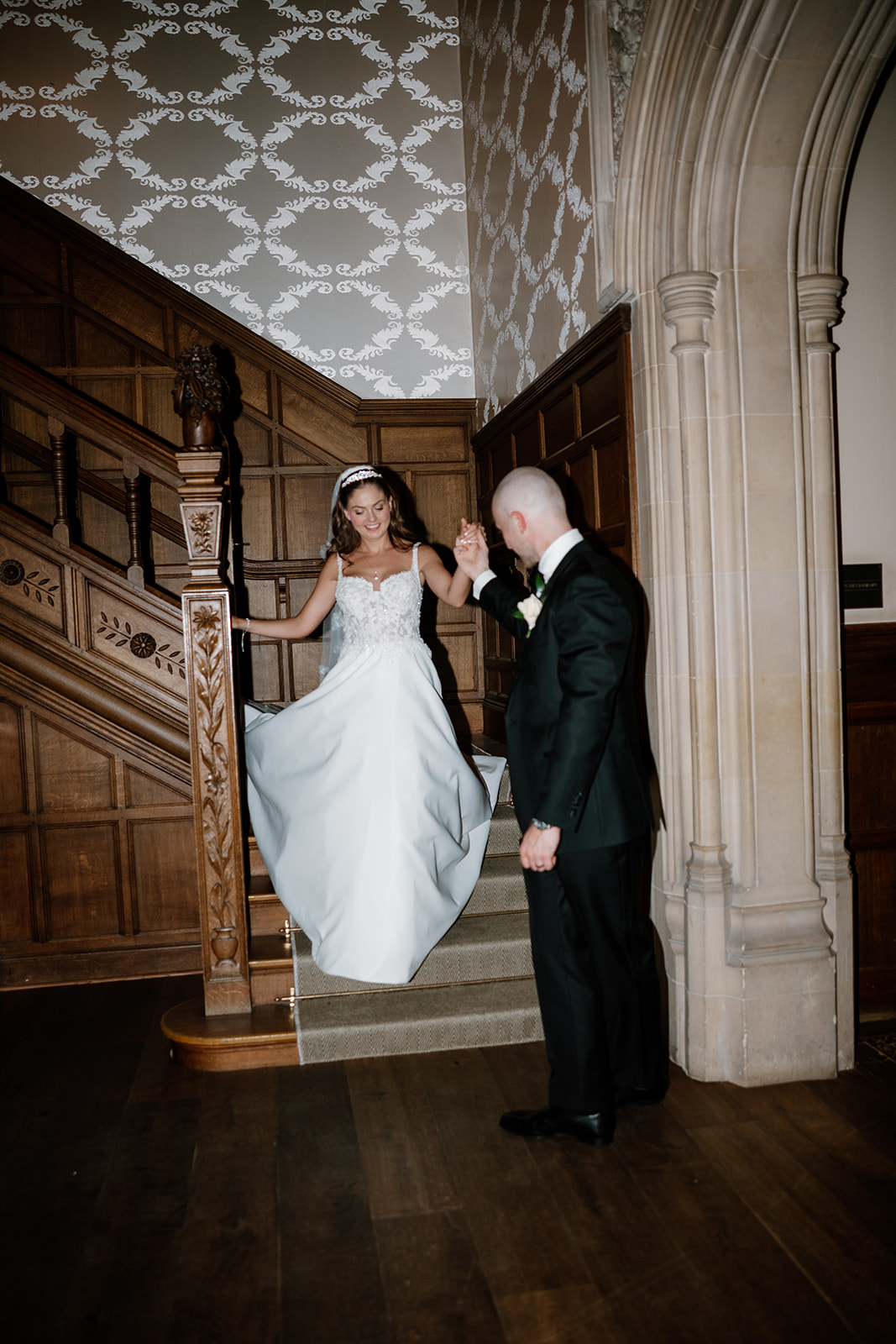 A bride in a white dress walks down wooden stairs at Hampton Manor, holding the hand of a man in a black suit inside a building with ornate wood panelling and an arched doorway.
