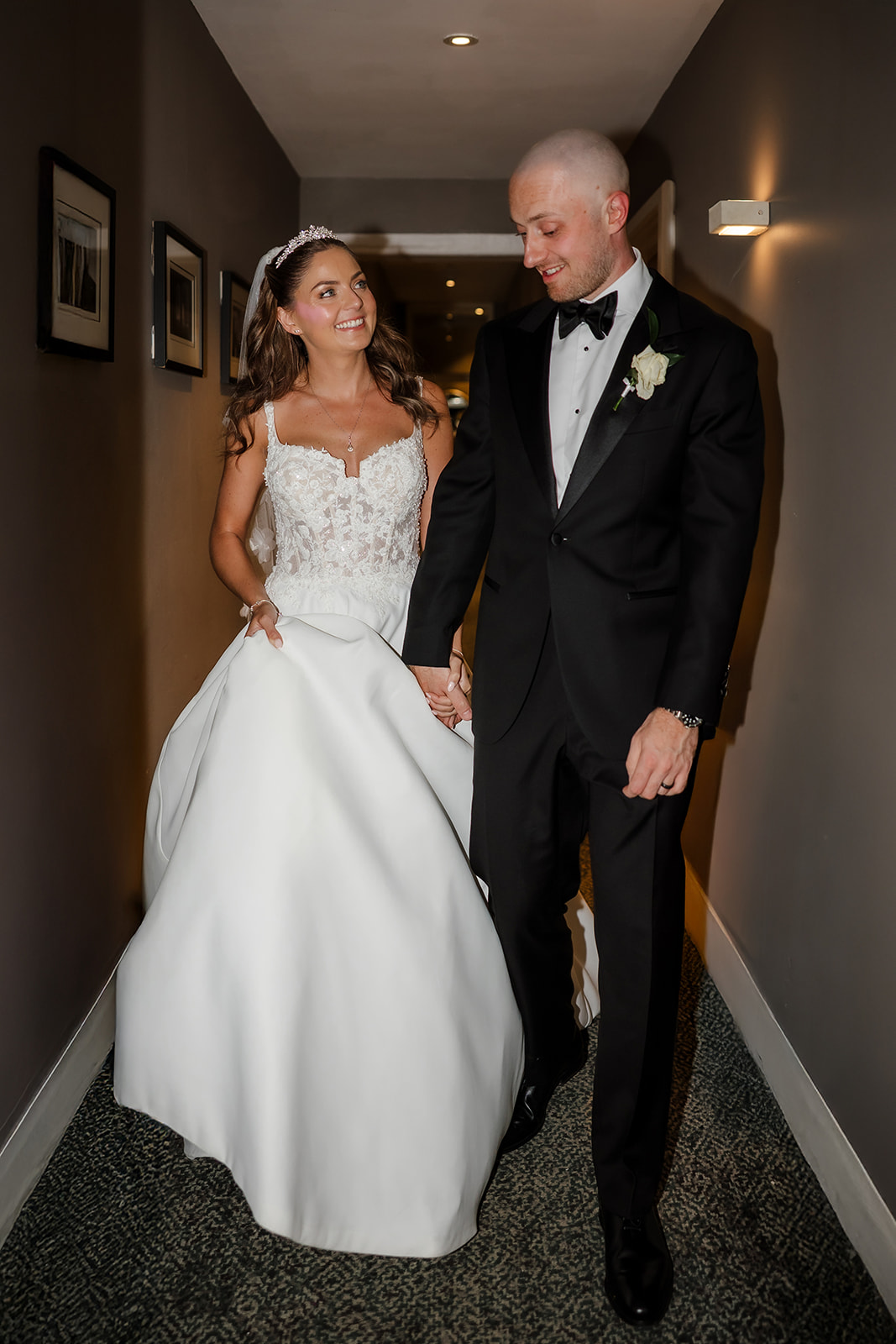 The bride and groom, with the bride in a white dress and the groom in a black dinner suit, walk hand in hand down a corridor, smiling at each other.