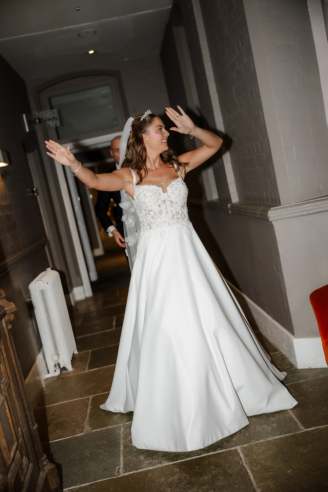A joyful bride in a white wedding dress stands in a corridor with her arms raised, smiling brightly, while another person in a suit stands supportively behind her.