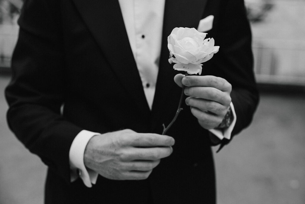 A person wearing a dinner jacket holds a single white rose in their hands. The image is in black and white.