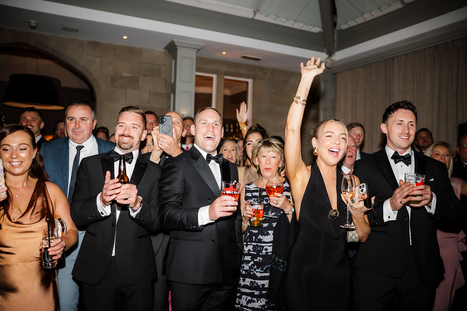 A group of well-dressed wedding guests at an indoor event hold drinks and smile, with one woman raising her hand enthusiastically, welcoming the couple.