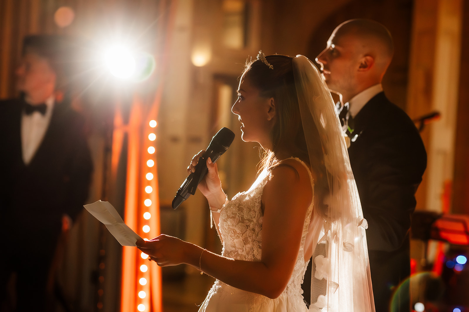 A bride giving a speech in her wedding dress holds a microphone and paper as she speaks, with the groom standing beside her in formal attire. Bright lights illuminate the scene.