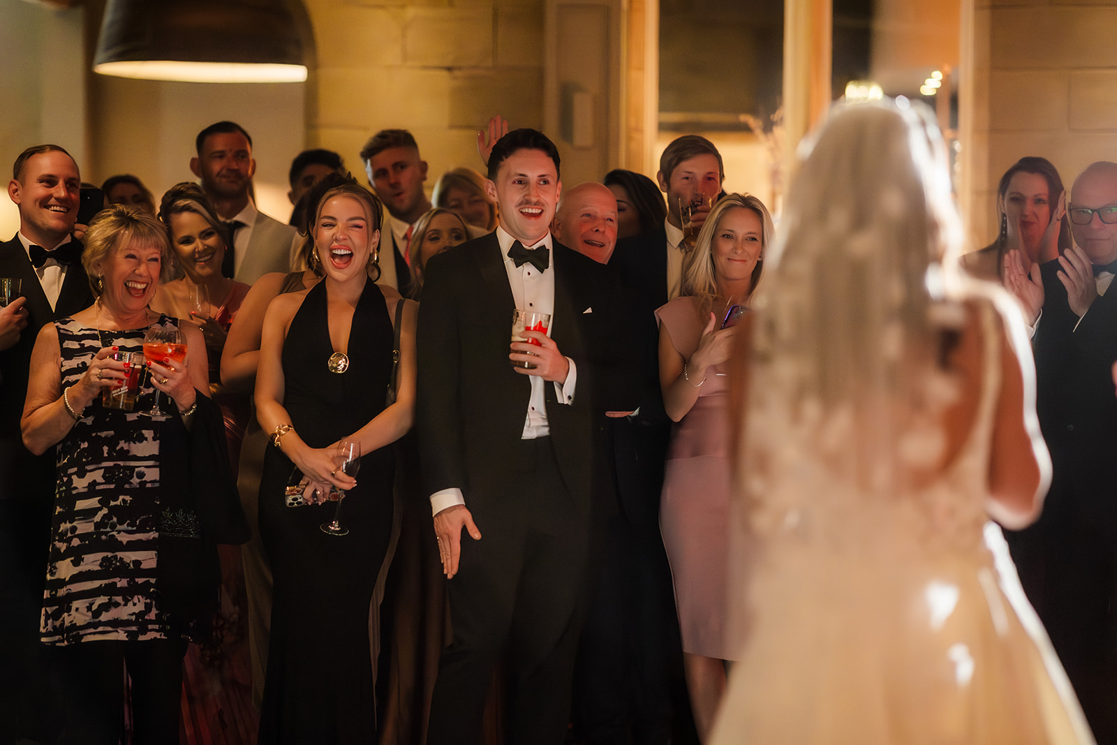 A group of well-dressed wedding guests, some holding drinks, smile and watch a bride in a wedding dress during a reception indoors.