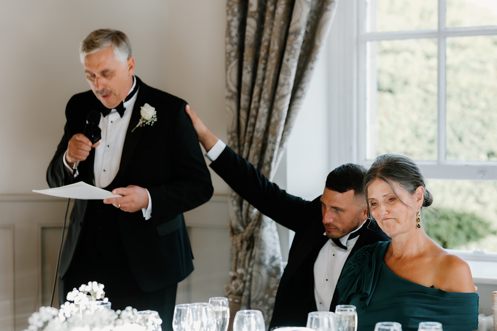 A man in a dinner jacket reads from a paper, delivering a heartfelt father of the bride speech at a formal event, as another man places a comforting hand on an emotional seated woman's shoulder.