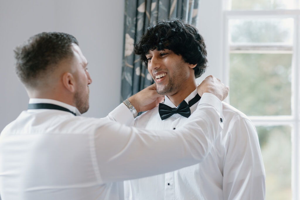 Two men in dress shirts and bow ties stand indoors; one adjusts the other's bow tie near a window with curtains.