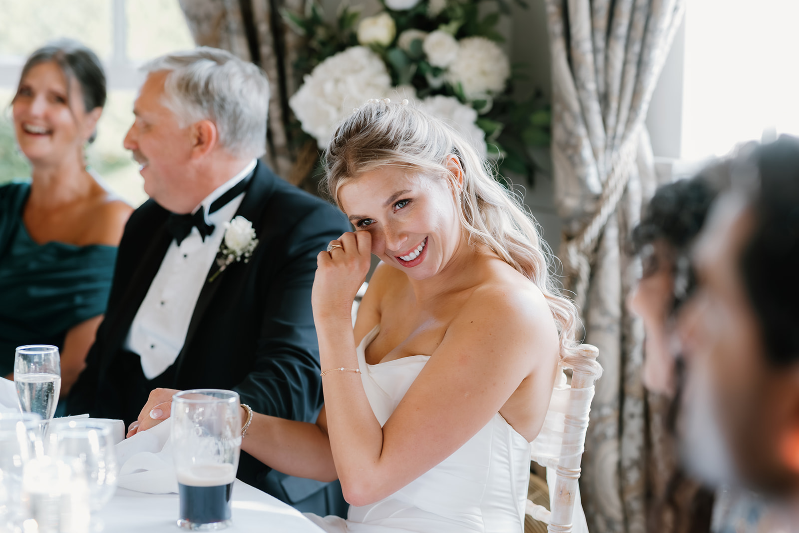 A bride in a white dress smiles and touches her face whilst seated at a table during a wedding reception, with other guests in formal attire nearby.