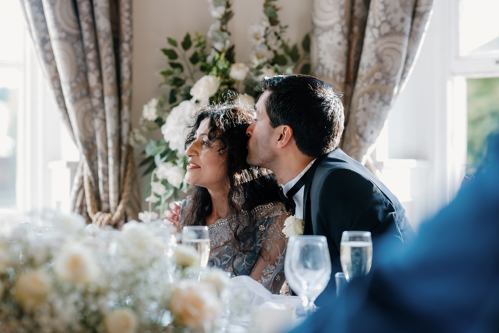 A man in a dinner jacket kisses a woman on the head as they sit at a table decorated with white flowers and glassware, with sunlight streaming through the windows behind them.