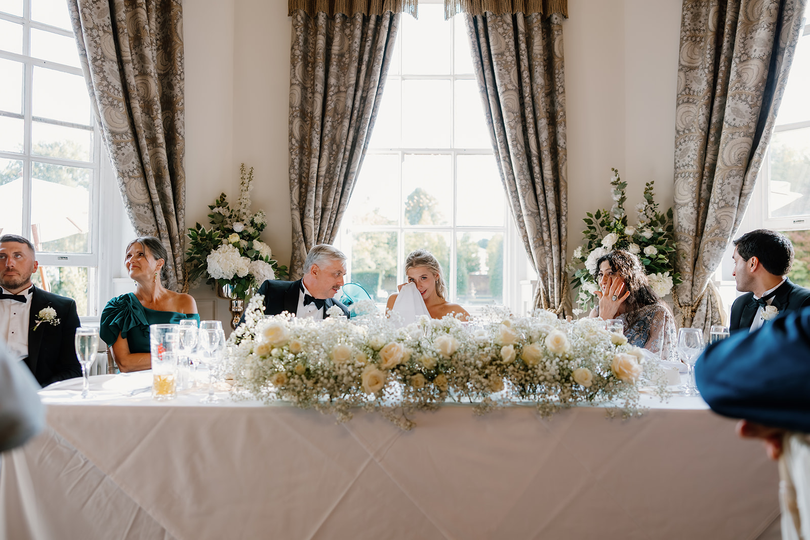 A wedding top table with six people dressed formally, floral centrepieces, and large windows with curtains in the background. The atmosphere appears emotional and celebratory.