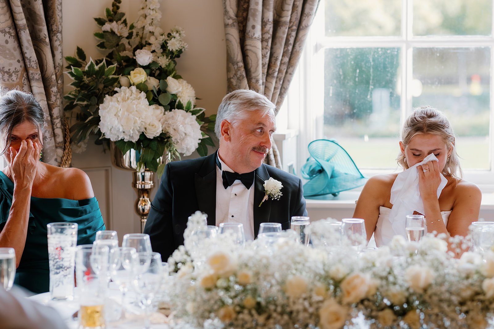 Three people sit at a decorated table; two women wipe their eyes while a man in a dinner jacket looks at them, smiling. Flowers and glassware are in the foreground, and a window is behind them.