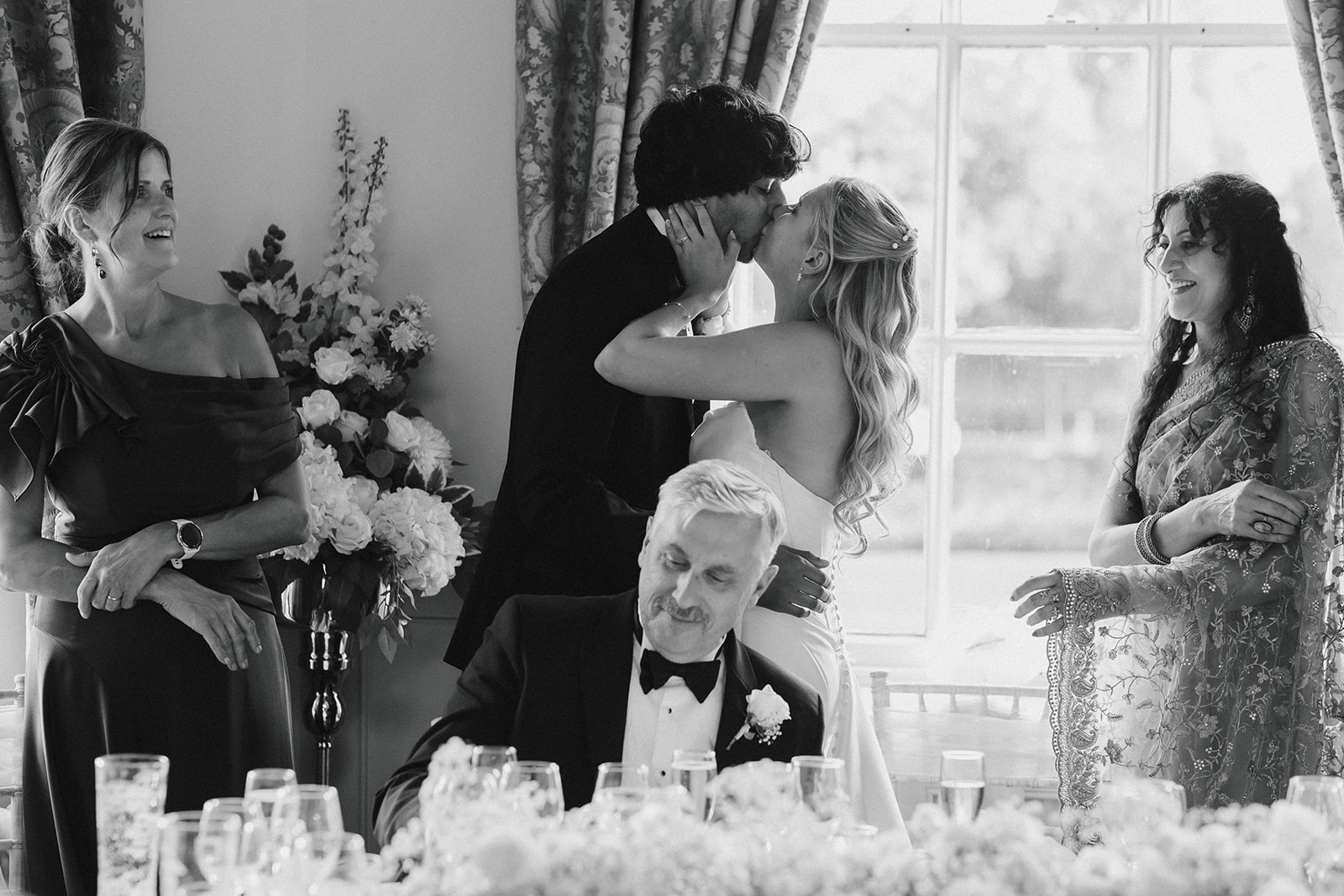 A bride and groom kiss whilst guests look on and smile at a formal indoor wedding reception with floral decorations.