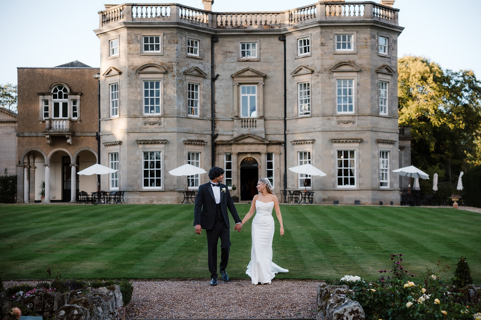 A bride and groom in formal attire walk hand in hand on a lawn in front of a large, elegant stone building with multiple windows and white parasols. Bourton Hall Wedding Photographer