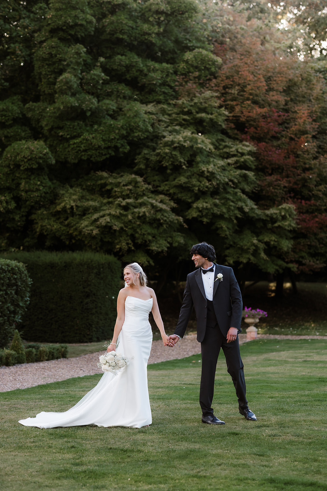 A bride in a white dress and a groom in a black tuxedo hold hands while standing on grass in a garden setting. Bourton Hall wedding photographer.