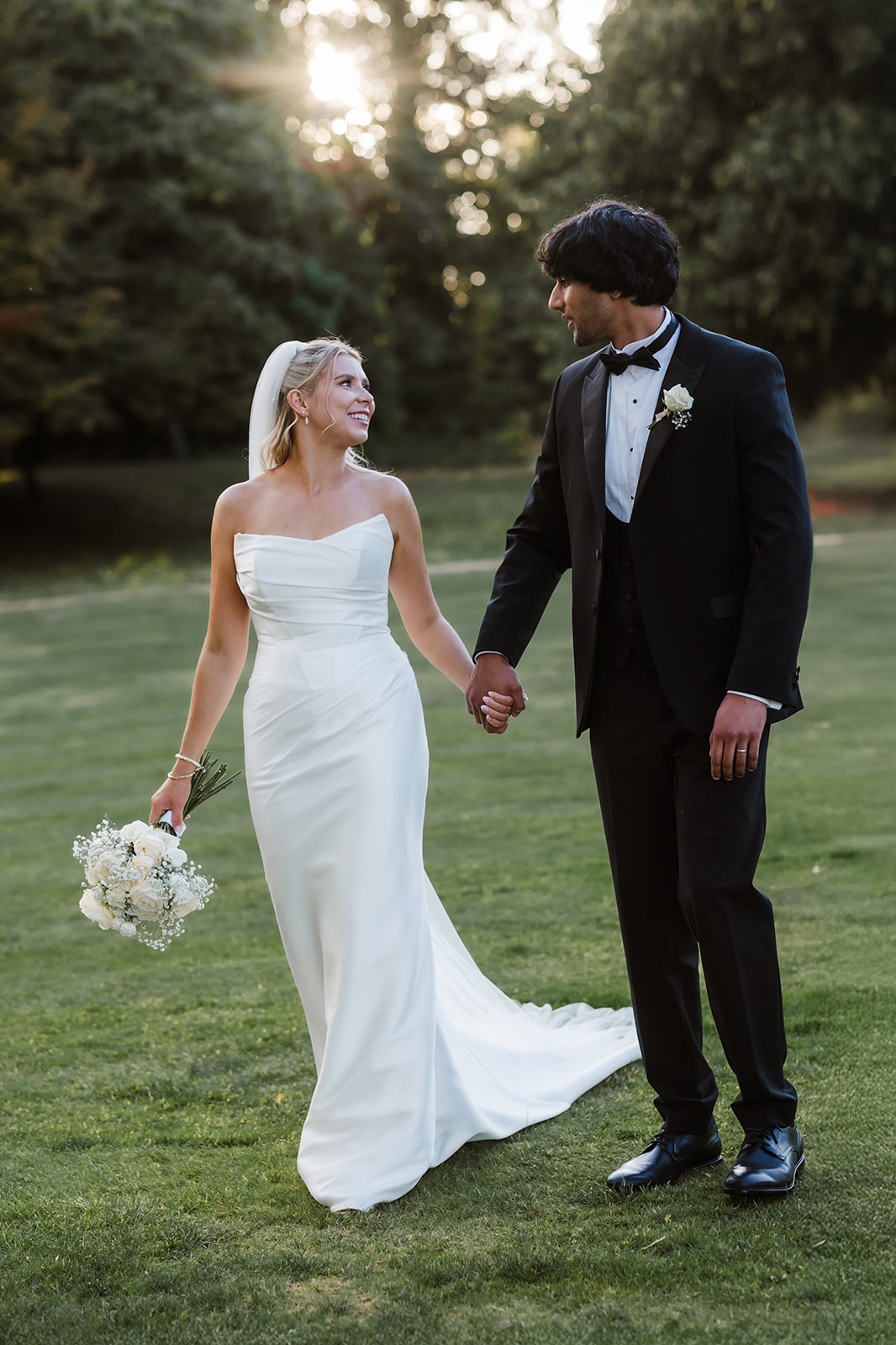 A bride in a white dress and a groom in a black dinner suit hold hands and walk on grass outdoors, both smiling at each other.
