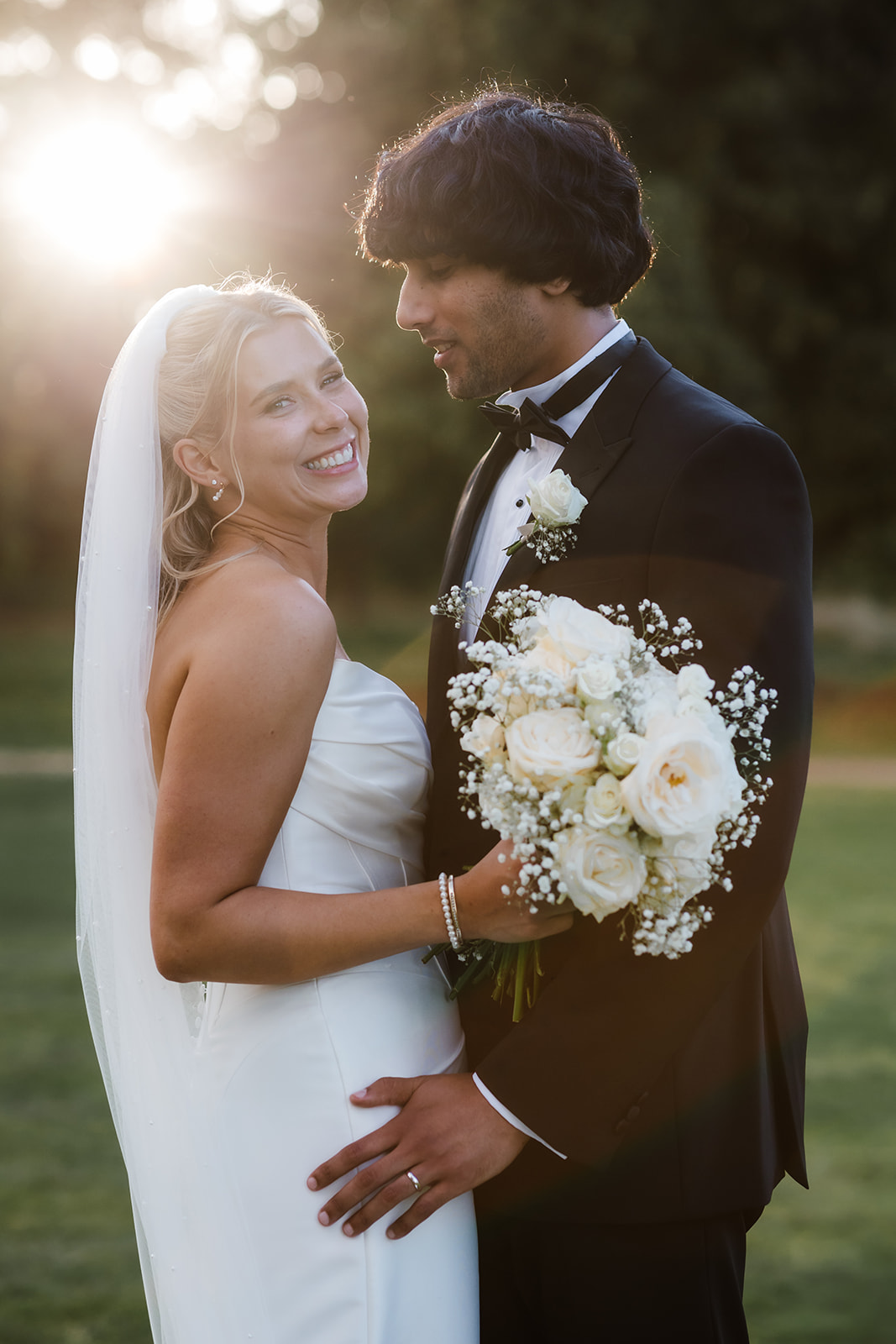 A bride in a white dress and veil holds a bouquet of white roses whilst smiling beside a groom in a black tuxedo, outdoors at golden hour.