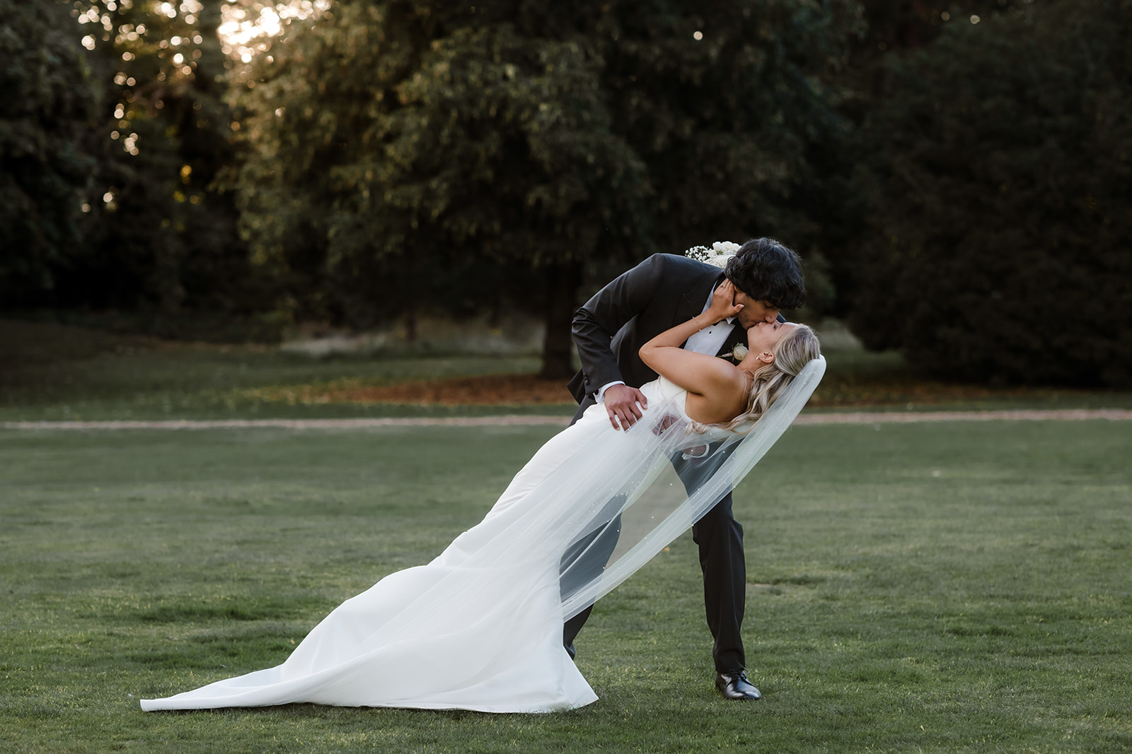 A groom in a black suit kisses a bride in a white dress and veil as he dips her on a grassy lawn with trees in the background.