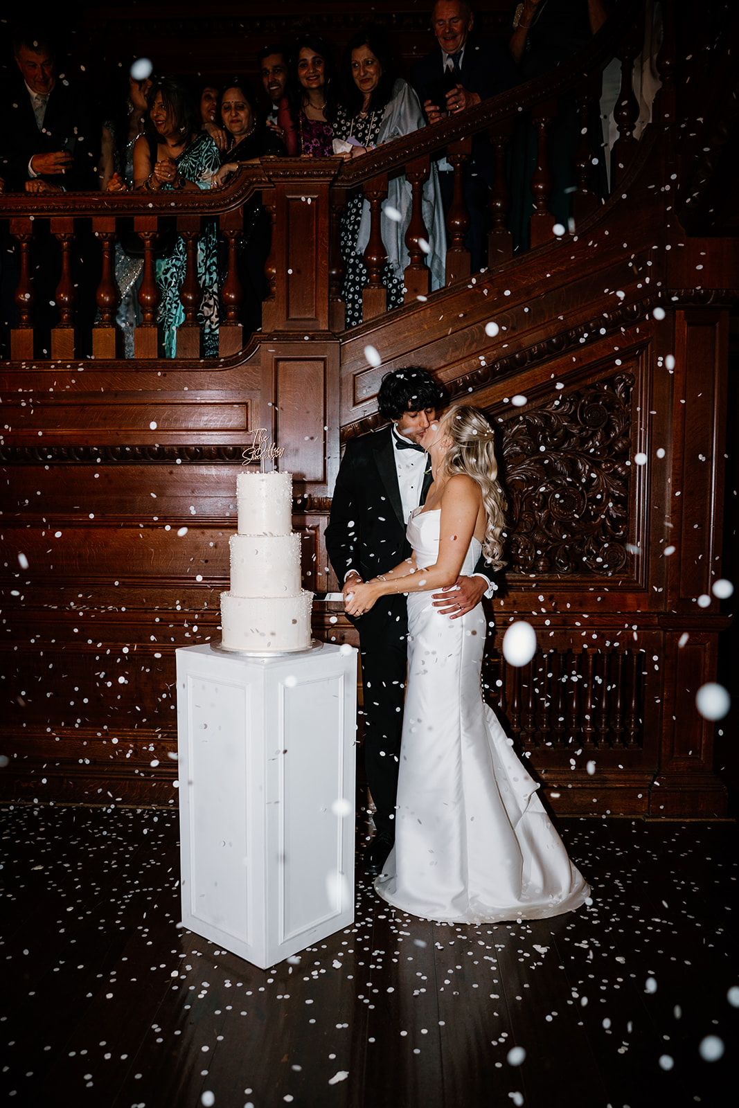 A bride and groom kiss in front of a wedding cake, surrounded by falling confetti, with guests watching from a wooden staircase behind them.