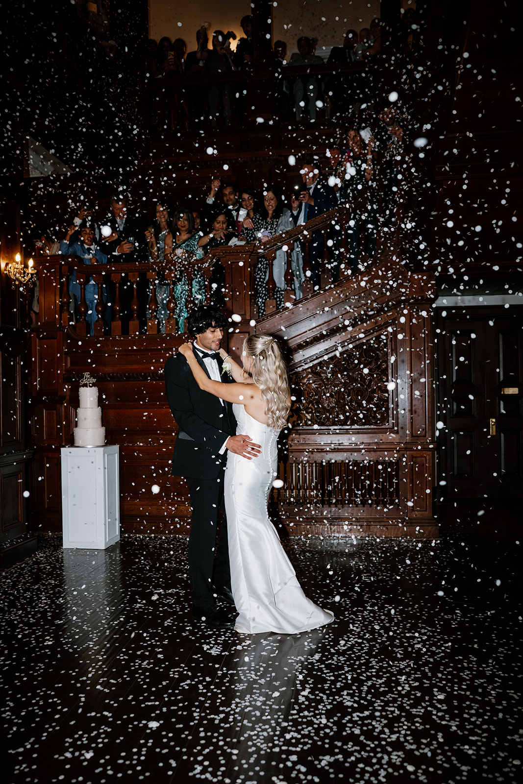 A bride and groom share a first dance on a dark wooden floor, surrounded by falling confetti, as guests watch from upstairs and the sides.