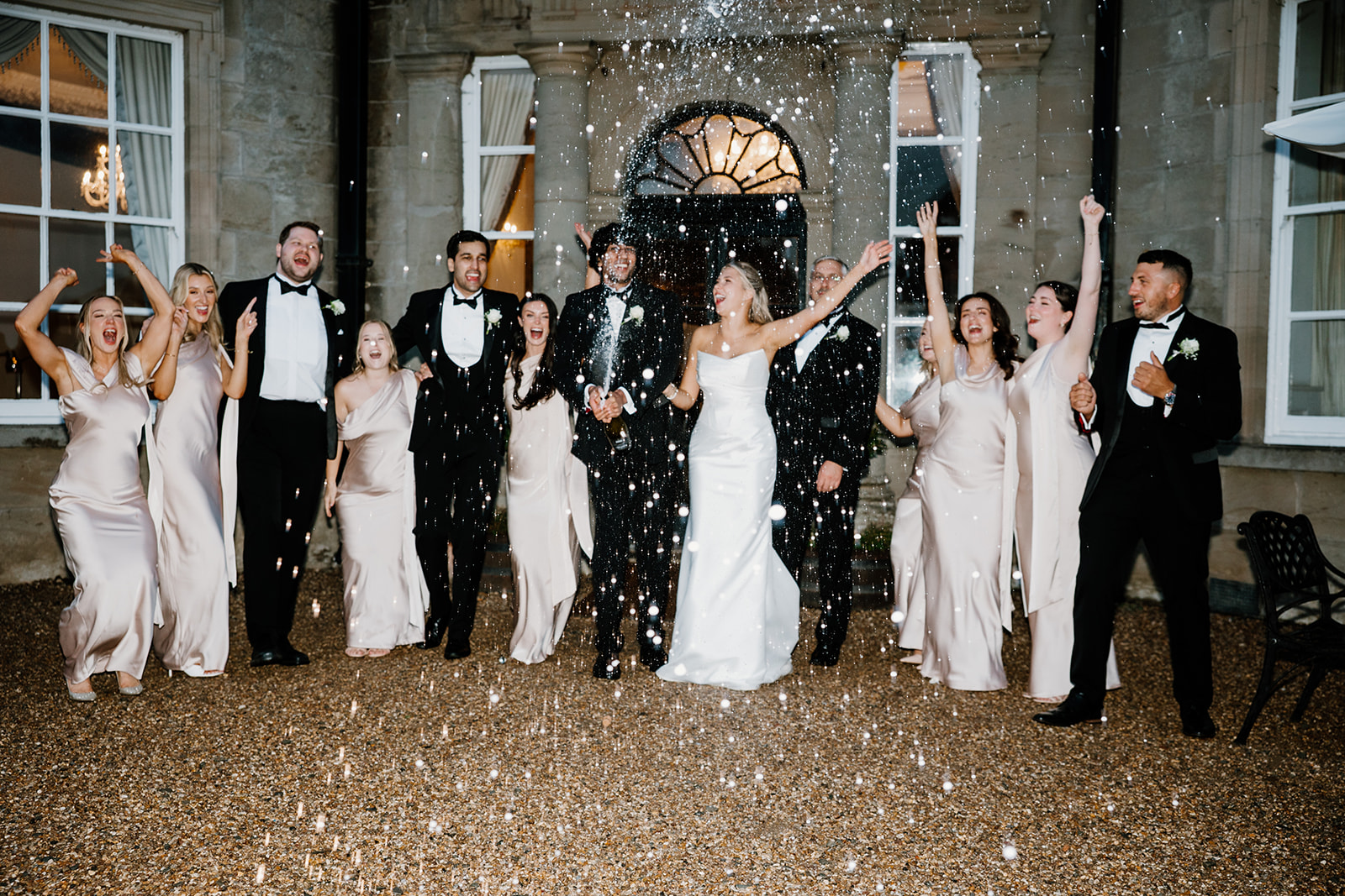 A wedding party in formal attire stands outside a building, celebrating and smiling as confetti or droplets fall around them.