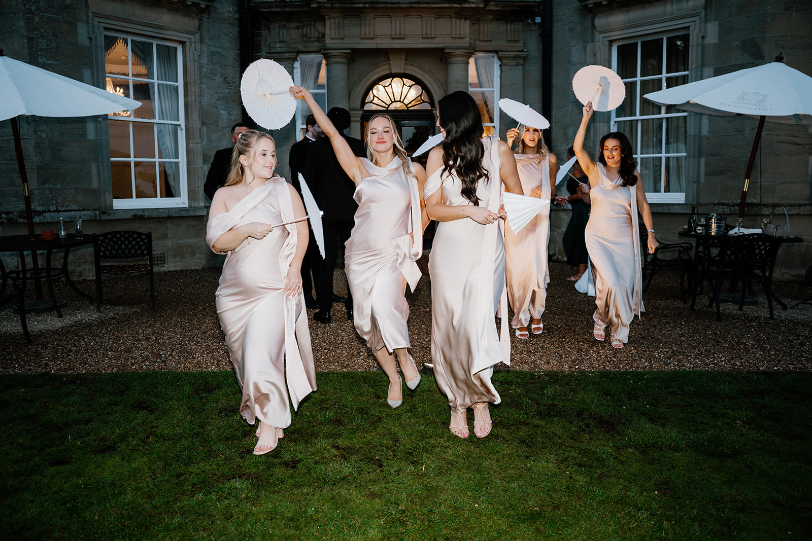 Bridesmaids in matching light-coloured dresses hold white parasols and walk or dance together on the grass outside a large stone building.