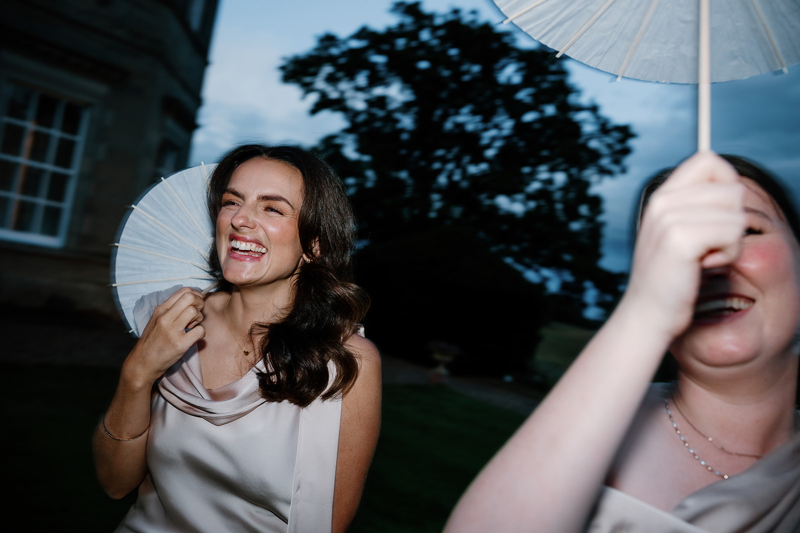 Two women in light-coloured dresses stand outside at dusk, one laughing and holding a paper parasol, the other partially obscured by her parasol.