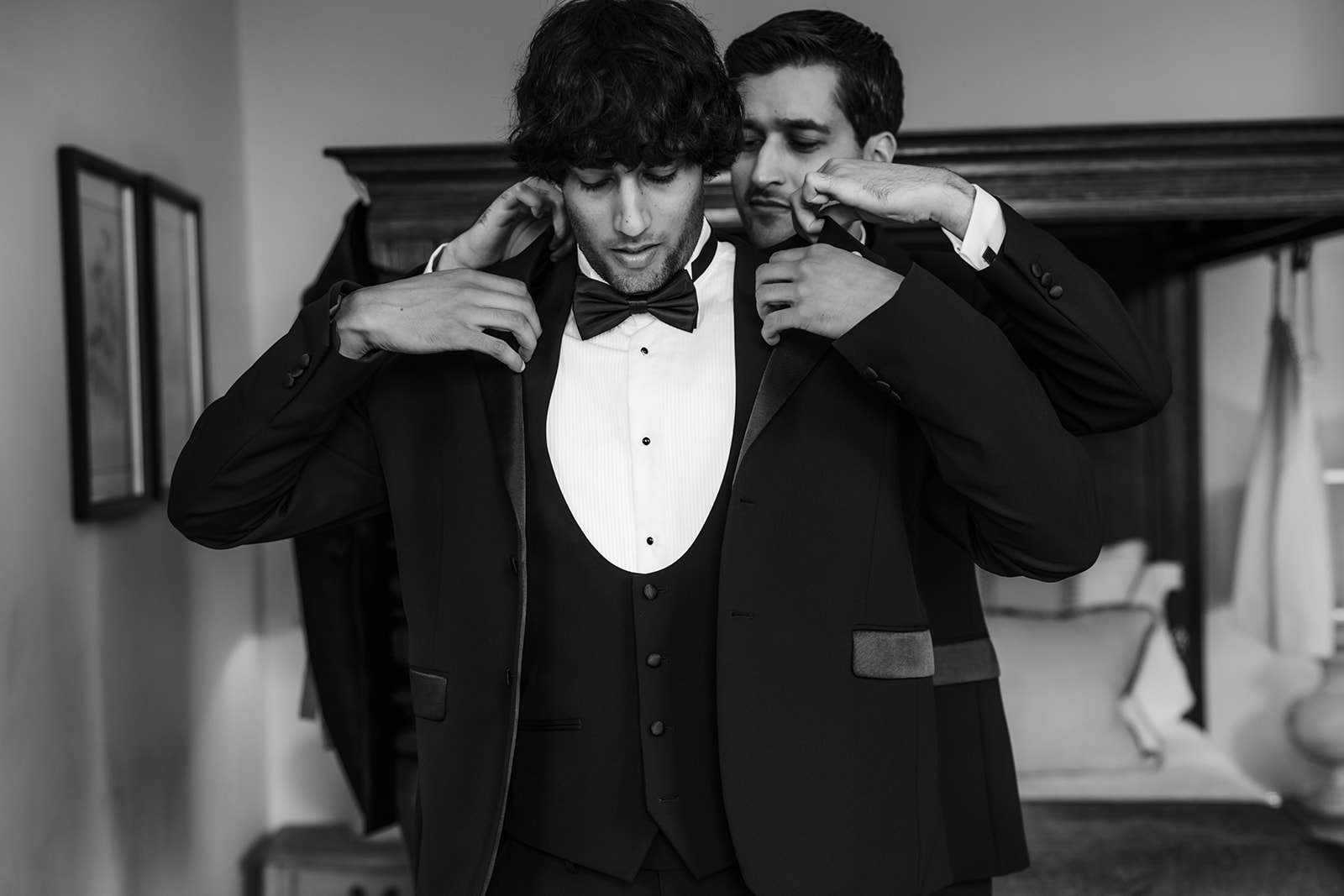 A groom and his brother, both in suits, stand in a bedroom as one helps the other adjust his jacket and bow tie, preparing for a formal occasion.