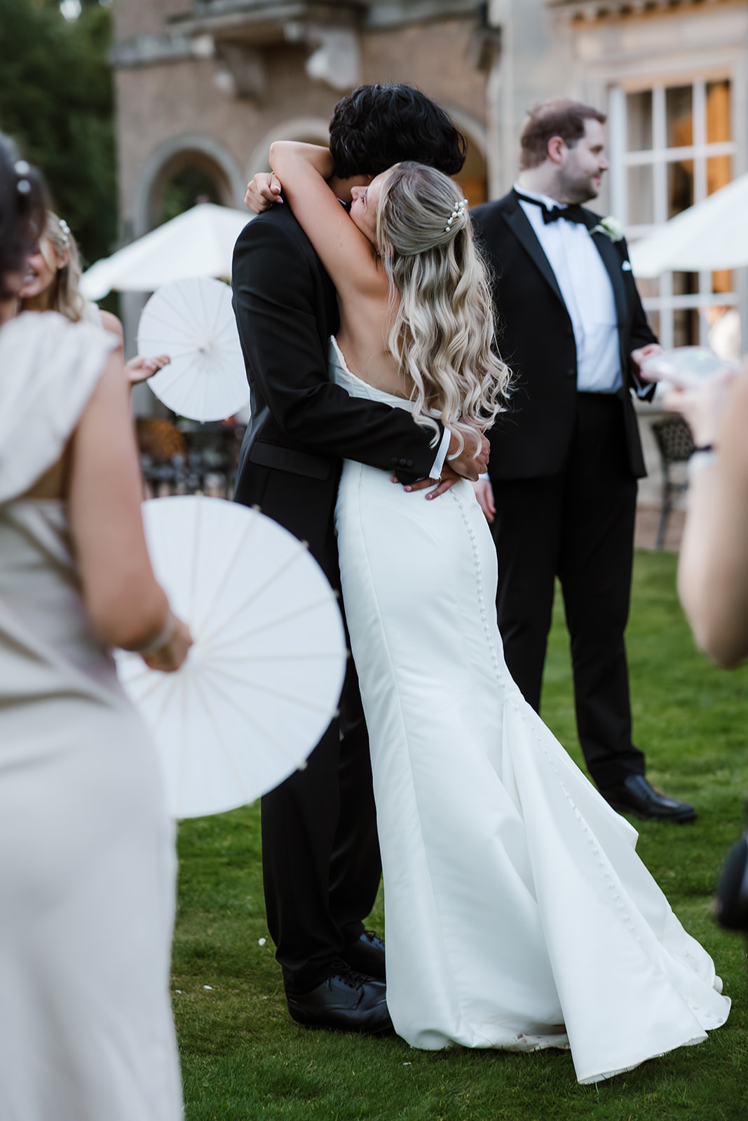 A bride and groom embrace and kiss outdoors at a wedding reception, with guests in formal attire and white parasols in the background.
