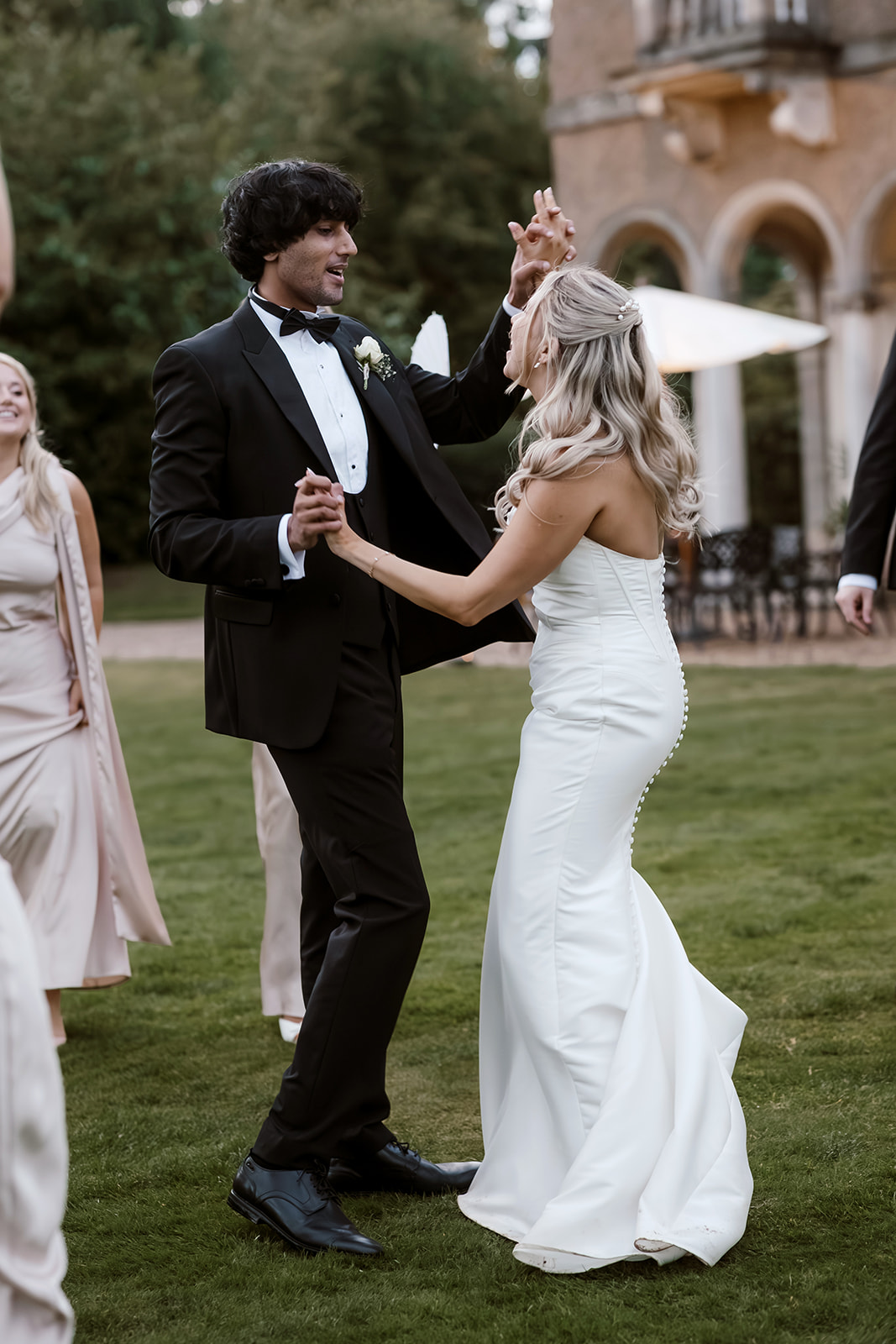 A bride and groom dance together outdoors on grass, surrounded by guests, with a stone building in the background.