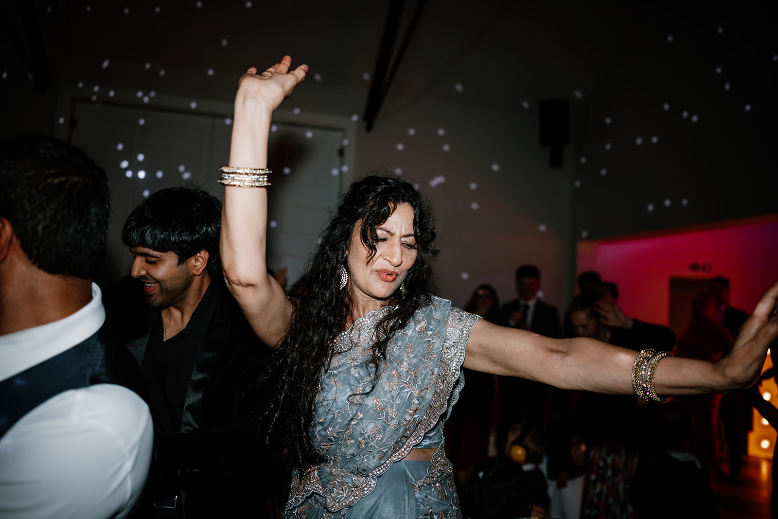 A woman in a blue sari with raised arm dances enthusiastically at an indoor event, surrounded by other people and dim, festive lighting.