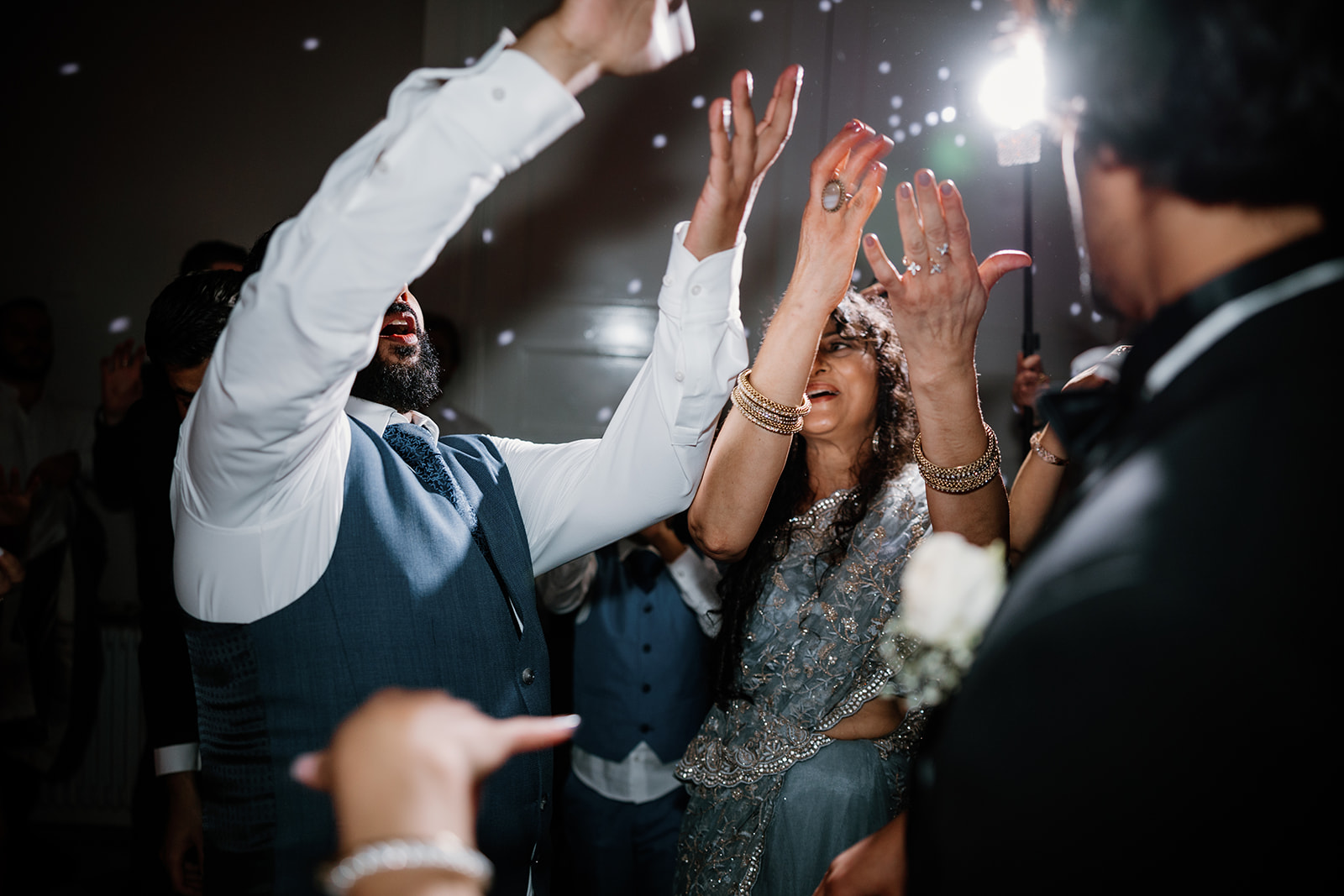 People dressed in formal attire dance and raise their hands at an indoor celebration, with bright lights and blurred motion in the background.