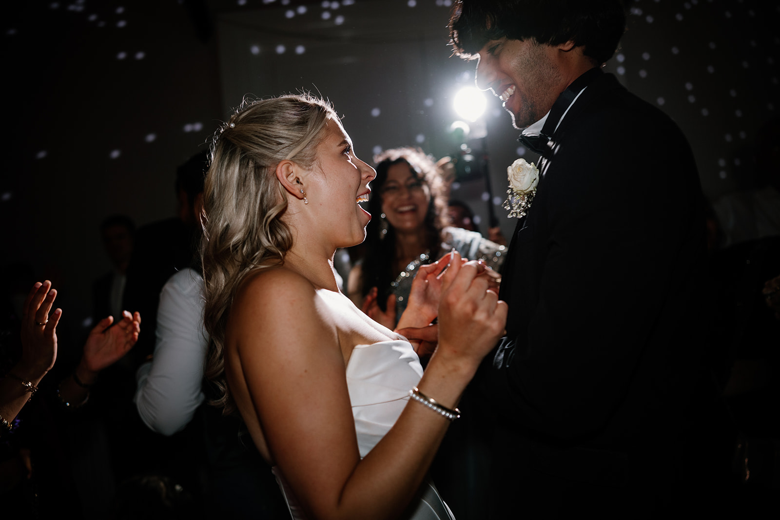 A bride and groom smile and hold hands while dancing at their wedding reception, surrounded by guests and soft lighting.