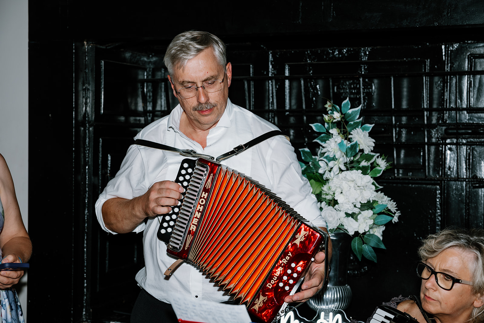 A man in a white shirt plays a red Hohner accordion indoors, with sheet music and a flower arrangement visible in the background.