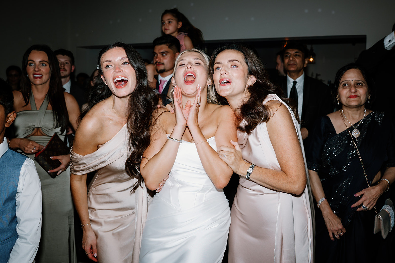 Three women in formal dresses, including a bride in white, stand close together smiling and singing at an indoor event, surrounded by other guests.