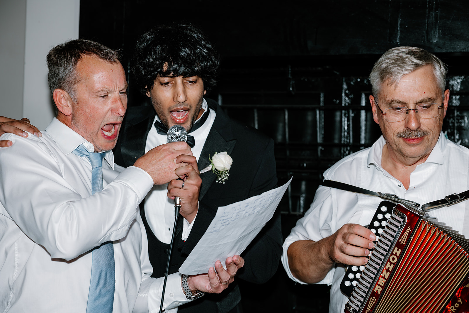Three men perform together; two sing into a microphone while the third plays an accordion. One man holds a sheet of paper, and all are dressed in formal shirts and jackets.