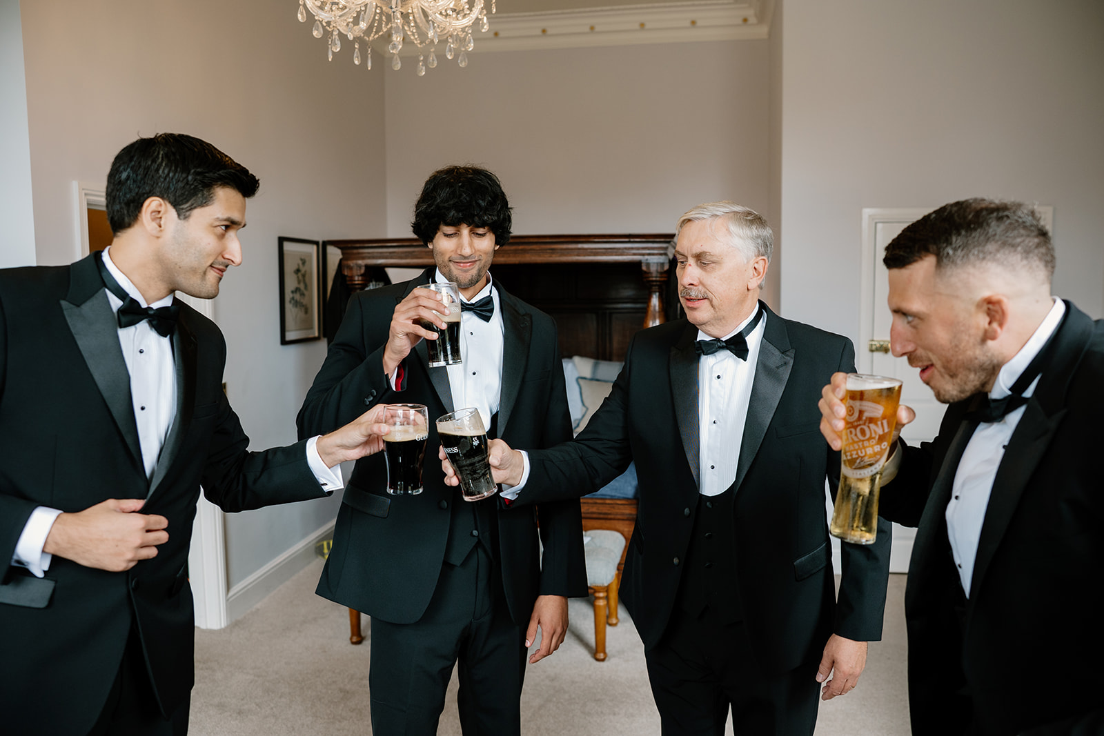 Four men in dinner jackets stand together indoors, holding drinks and laughing—groom and groomsman celebrating or toasting in a well-lit room with a chandelier and a bed in the background.