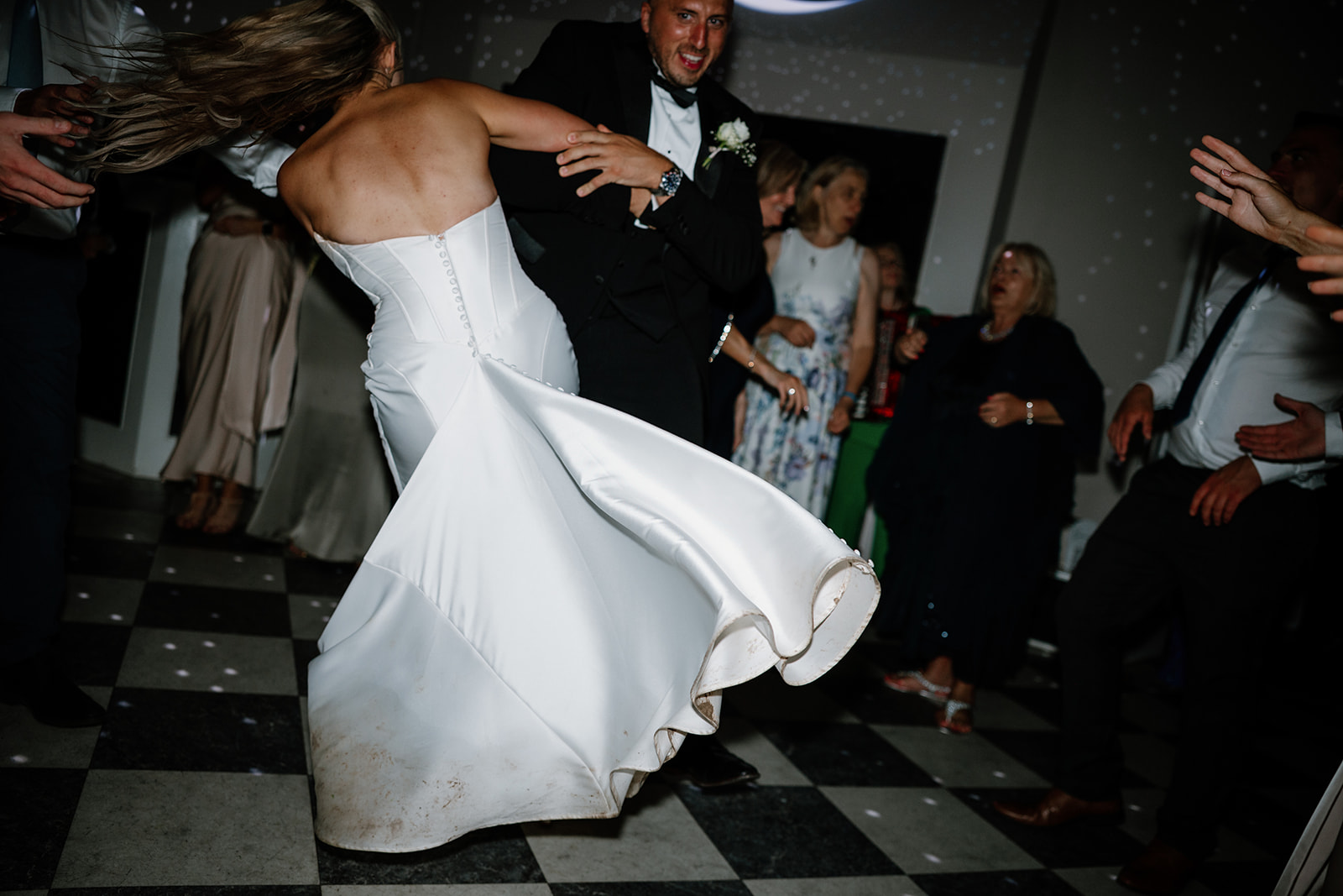 A bride in a white dress twirls with a man in a suit on a chequered dance floor, surrounded by guests at a wedding reception.
