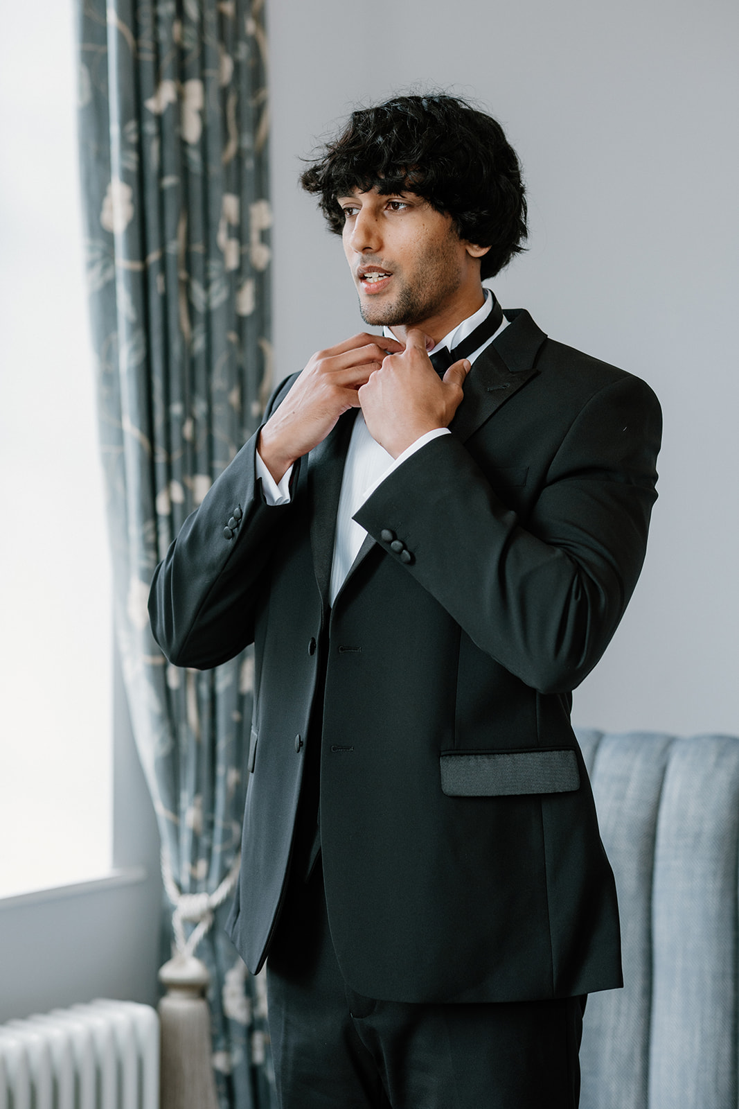 A groom in a black suit adjusts his bow tie whilst standing indoors near a window with patterned curtains.
