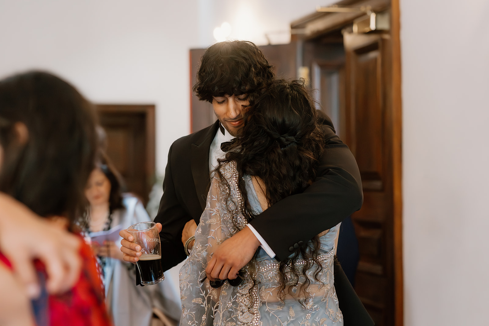 The groom and his mother share a heartfelt hug, both dressed elegantly, as he holds a drink in one hand at a festive indoor event.