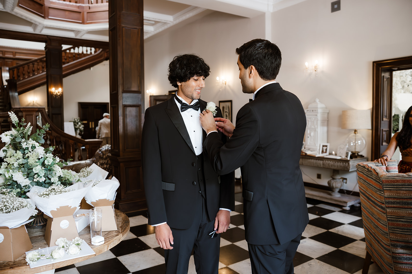 Two men in dinner jackets stand in a formal room; one is pinning a buttonhole on the other's jacket. Floral arrangements and wedding decor are visible on a nearby table.