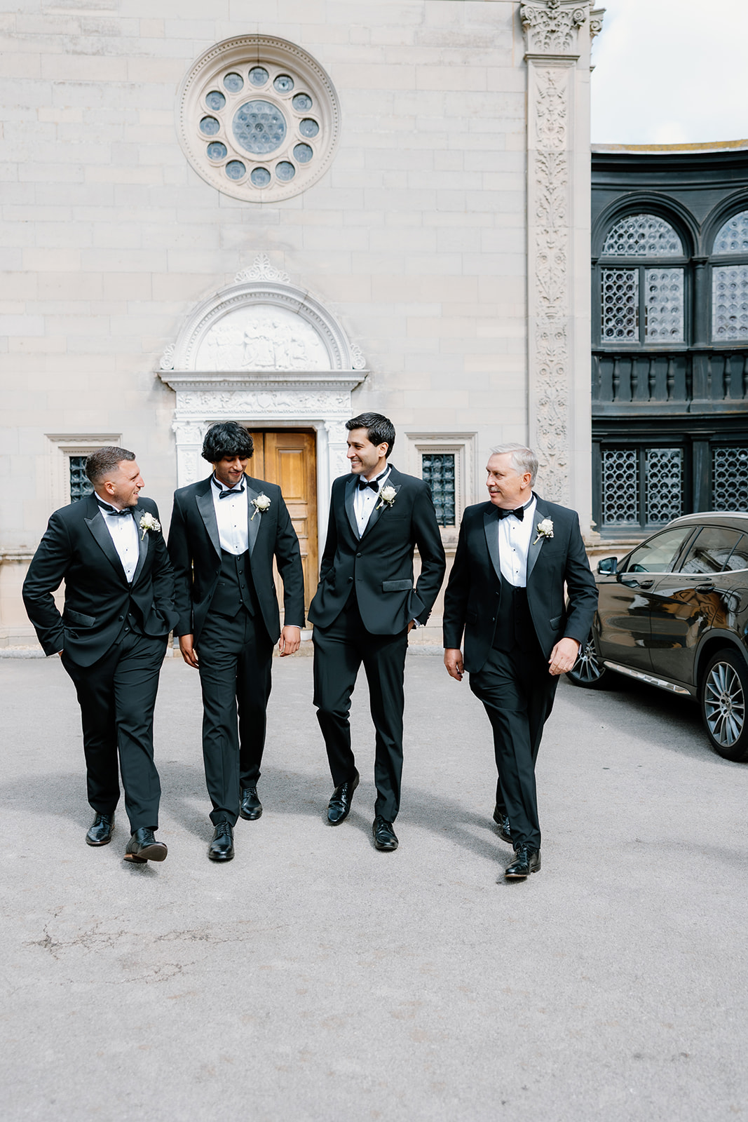 Four men in dinner jackets and buttonholes walk together outside a light stone building with ornate windows and a parked black car nearby.