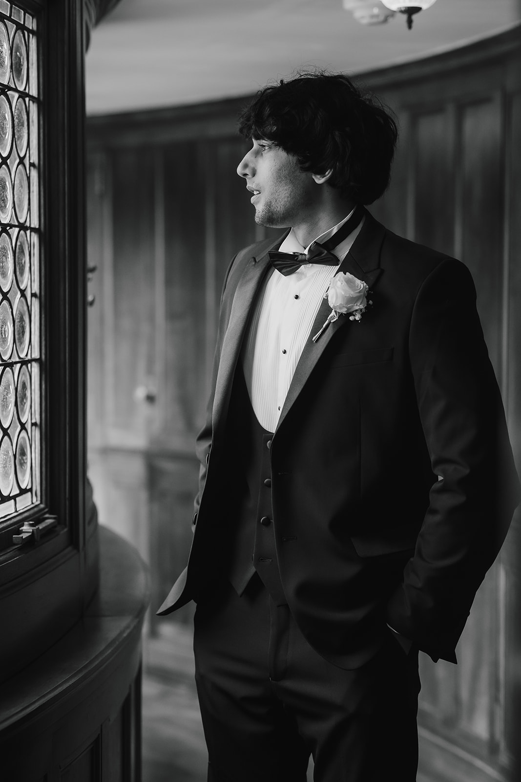 The groom, in a tuxedo with a buttonhole, stands indoors by a leaded glass window, looking outside with one hand in his pocket.
